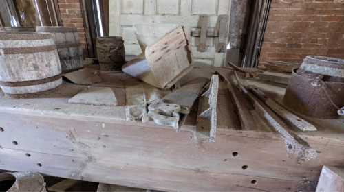 A cluttered wooden table with various tools, barrels, and debris in a rustic, dimly lit space.