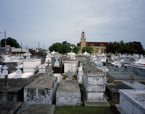 A cemetery filled with tombstones and mausoleums, with a church in the background under a cloudy sky.