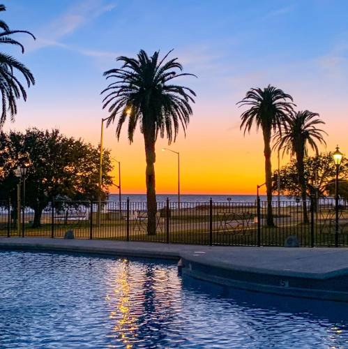 A serene sunset over a pool, framed by palm trees and a view of the ocean in the background.