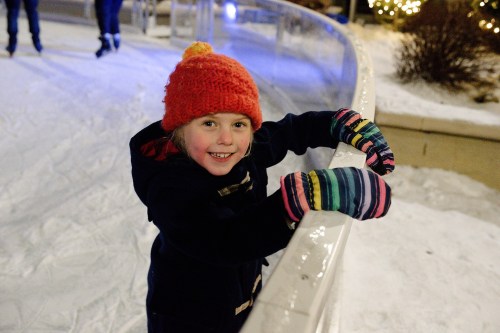 A smiling child in a colorful hat and mittens leans on a railing at an ice skating rink, surrounded by snow.