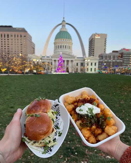 Two hands hold a plate of sliders and loaded tater tots, with a city skyline and arch in the background.