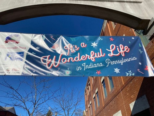 Banner reading "It's a Wonderful Life in Indiana, Pennsylvania" against a blue sky, with festive snowflakes and decorations.