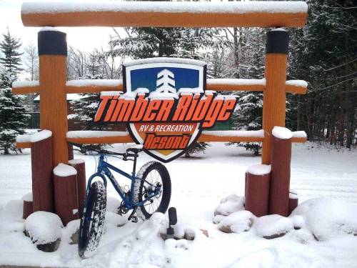 A snowy scene featuring a sign for Timber Ridge RV & Recreation Resort, with a blue bike leaning against it.