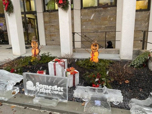 Two snowman decorations and ice sculptures with gifts in front of a building, surrounded by holiday greenery and snow.