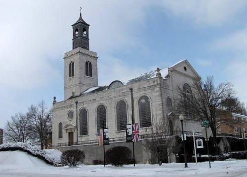 Historic church with a clock tower, surrounded by snow and trees, under a cloudy sky. Flags are displayed outside.
