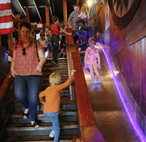 A busy indoor space with a slide; a woman walks down stairs, while a child plays and another child slides down.