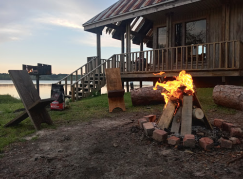 A cozy outdoor scene featuring a fire pit with flames, wooden chairs, and a rustic cabin by the water at sunset.