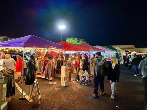 A vibrant night market with colorful tents, shoppers browsing clothing, and festive lights illuminating the scene.