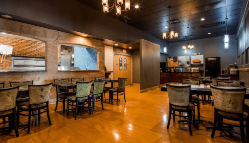 Interior of a modern restaurant featuring wooden floors, tables, and chairs, with warm lighting and a brick wall.