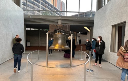 A large bell displayed indoors, surrounded by visitors wearing masks in a modern architectural space.