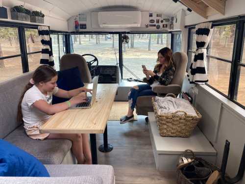 A girl uses a laptop at a table while a woman sits nearby, both inside a cozy bus converted into a living space.