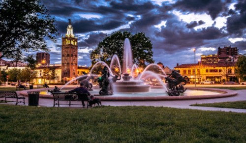 A fountain in a park at dusk, surrounded by benches, with a city skyline and dramatic clouds in the background.