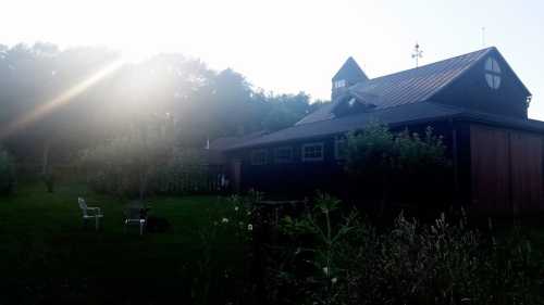 A rustic black barn surrounded by greenery, with sunlight shining through trees in the background.