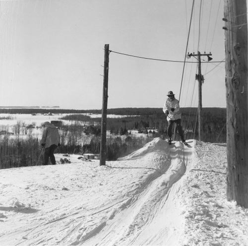 A skier descends a snowy slope near power lines, while another person walks in the background. Winter landscape visible.