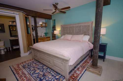 Cozy bedroom with a queen bed, teal walls, wooden accents, and a patterned rug. Includes a kitchenette and bathroom area.