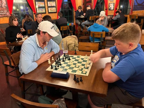 A man and a boy play chess at a busy café, with other patrons in the background. The chessboard is set up for a match.
