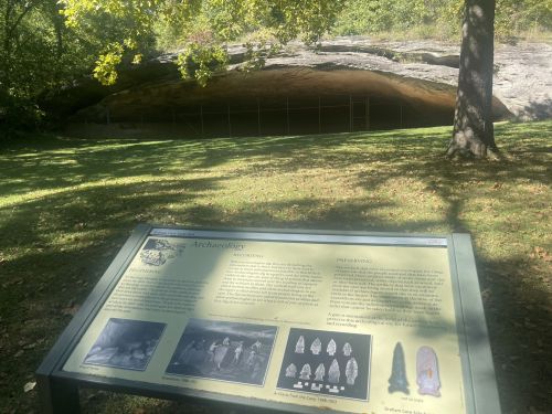 A grassy area with a large rock overhang and an informational sign about archaeology in the foreground.