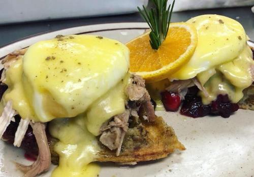 Two eggs Benedict with hollandaise sauce on a bed of pulled meat and cranberry, garnished with an orange slice and rosemary.
