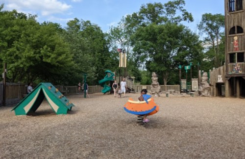 A playground with a child on a spinning toy, slides, climbing structures, and trees in the background.