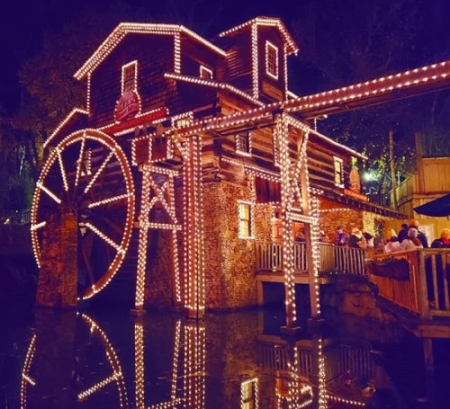 A beautifully lit watermill at night, adorned with festive lights reflecting in the water below.