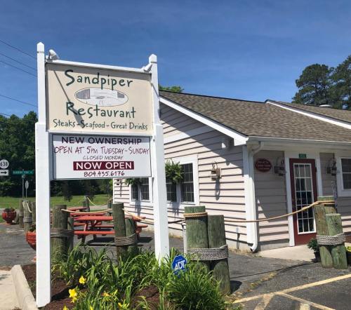 Sign for Sandpiper Restaurant, featuring "New Ownership" and hours, with a cozy building and outdoor seating visible.