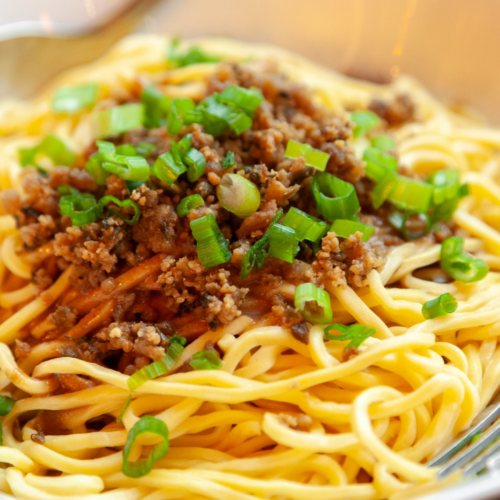 A close-up of noodles topped with ground meat and chopped green onions in a bowl.