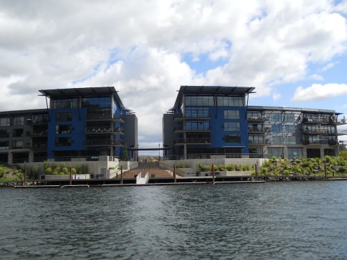 Modern waterfront apartments with blue accents, surrounded by greenery and a calm waterway under a cloudy sky.