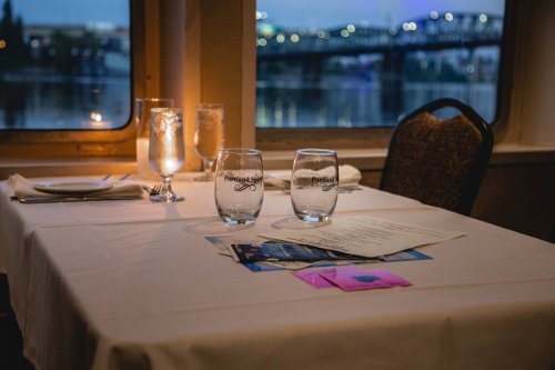 A dining table set for two by a window, featuring glasses, a candle, and a menu, with a view of a bridge outside.