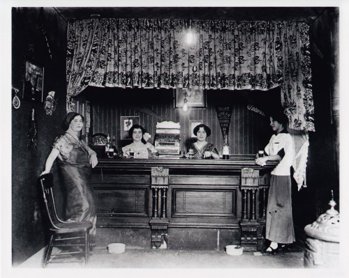 A vintage black-and-white photo of four women behind a bar, with decorative curtains and various items on the counter.