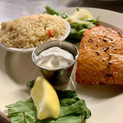 A plate featuring grilled salmon, rice, a lemon wedge, and a small bowl of sauce, with greens in the background.