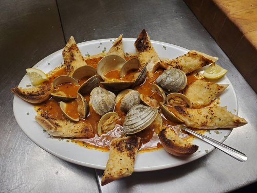 A platter of clams in a spicy sauce, surrounded by toasted bread and garnished with lemon wedges.