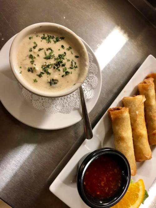 A bowl of creamy soup garnished with herbs, served with spring rolls and a side of dipping sauce on a plate.