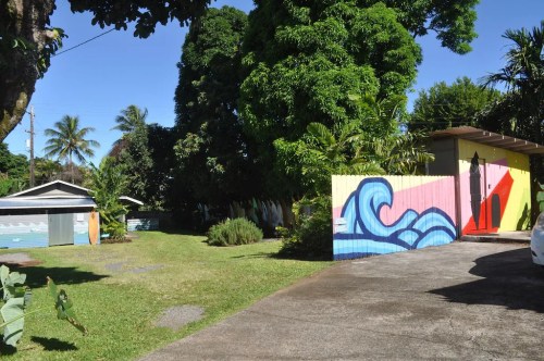 A colorful mural on a fence beside a driveway, with lush greenery and palm trees in a sunny residential area.
