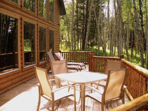 A wooden deck with a table and chairs, surrounded by trees and sunlight filtering through the leaves.