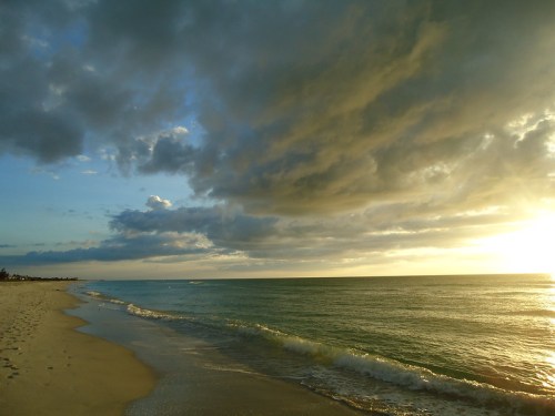A serene beach scene at sunset, with gentle waves and dramatic clouds in the sky.