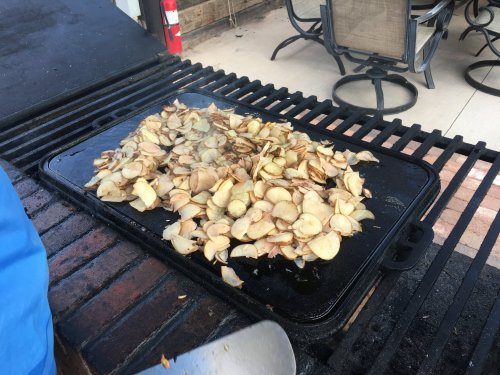 Sliced potatoes cooking on a large griddle outdoors, with a patio and chairs in the background.