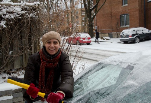A woman in a winter coat and hat scrapes snow off a car, smiling in a snowy urban setting.