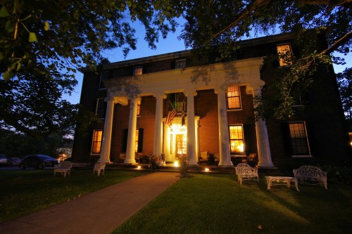 A historic brick building with white columns, illuminated at dusk, featuring an American flag and surrounded by greenery.