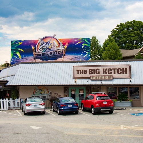 Exterior of The Big Ketch Saltwater Grill with a colorful sign and parked cars in front.