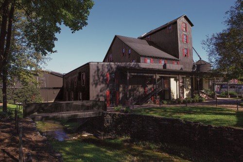 Historic building with red shutters, surrounded by greenery and a small stream, under a clear blue sky.