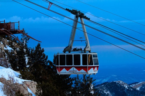 A ski gondola suspended over snowy mountains, with a blue sky in the background.