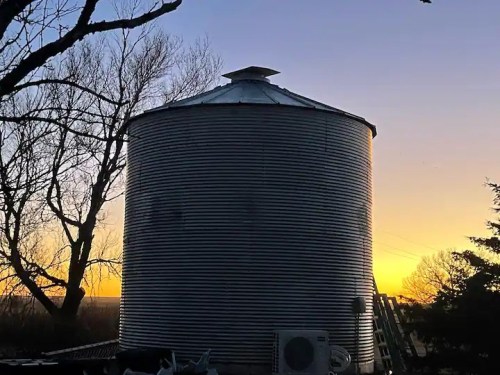 A metal grain silo silhouetted against a colorful sunset, with bare trees in the foreground.