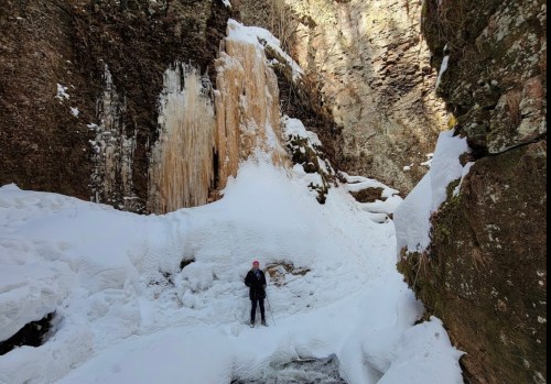 A person stands in a snowy canyon near a frozen waterfall, surrounded by rocky cliffs and ice formations.