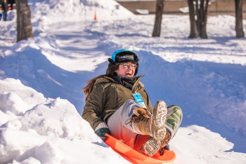 A person joyfully sledding down a snowy hill, wearing winter gear and a blue helmet, with snow all around.