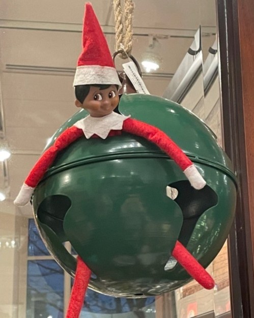 An elf doll with a red hat and arms sitting inside a green ornament, hanging from a string.