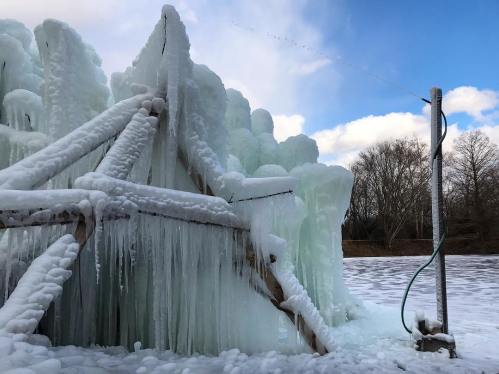 A frozen structure with icicles, surrounded by snow and a blue sky with clouds in the background.