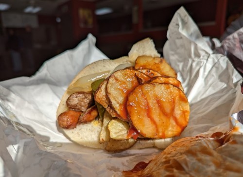 A sandwich with sausage, pickles, and fried potato slices, drizzled with sauce, wrapped in paper.