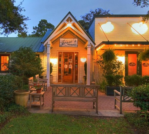 A charming restaurant exterior with warm lighting, benches, and greenery, inviting guests to enter.