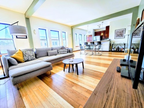 Bright living room with a gray sofa, wooden floors, and an open kitchen in the background. Cozy and modern atmosphere.