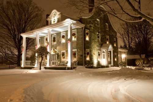 A snow-covered historic house illuminated at night, decorated with wreaths and holiday lights.